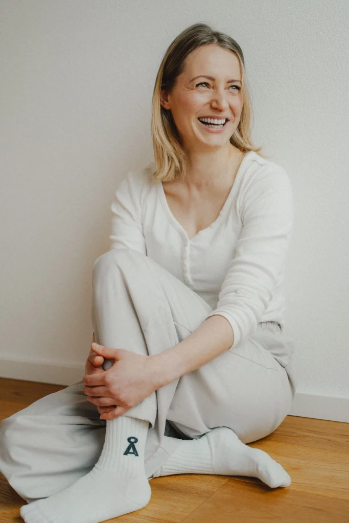 Hebamme Isabel Braun sitzt entspannt auf einem Holzboden an einer weißen Wand. Sie hat blondes Haar und trägt ein weißes Langarmshirt, eine helle Jogginghose und weiße Socken mit einem dunkelblauen Symbol darauf.