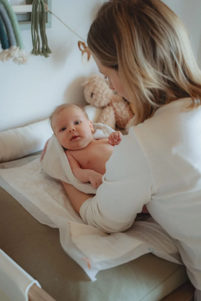 Die Hebamme Isabel Braun wickelt ein Neugeborenes auf einem Wickeltisch. Das Baby ist in ein Handtuch gewickelt und blickt ruhig in die Kamera. Im Hintergrund sind ein Kuscheltier und Dekorationen an der Wand zu sehen.