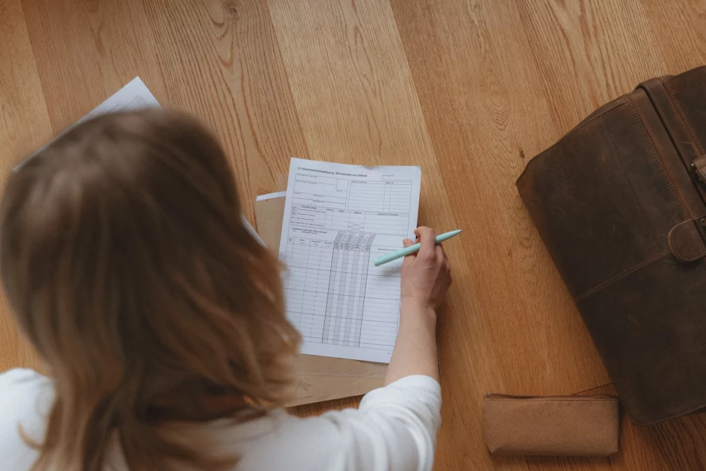 Die Hebamme Isabel Braun sitzt auf einem Holzboden und füllt ein Formular aus, welcher zur Unterstützung in der Schwangerschaft dient. Neben ihr liegen eine braune Ledertasche und ein Federmäppchen.