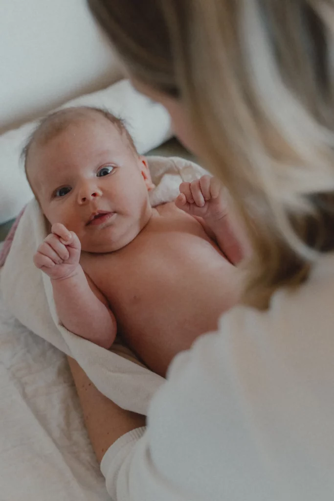Ein nacktes Baby liegt in einem Handtuch auf dem Arm der Hebamme Isabel Braun und schaut mit großen, wachen Augen nach oben. Isabel ist nur teilweise von hinten zu sehen und hält das Baby fürs Wickeln. Die Szene wirkt ruhig und liebevoll.
